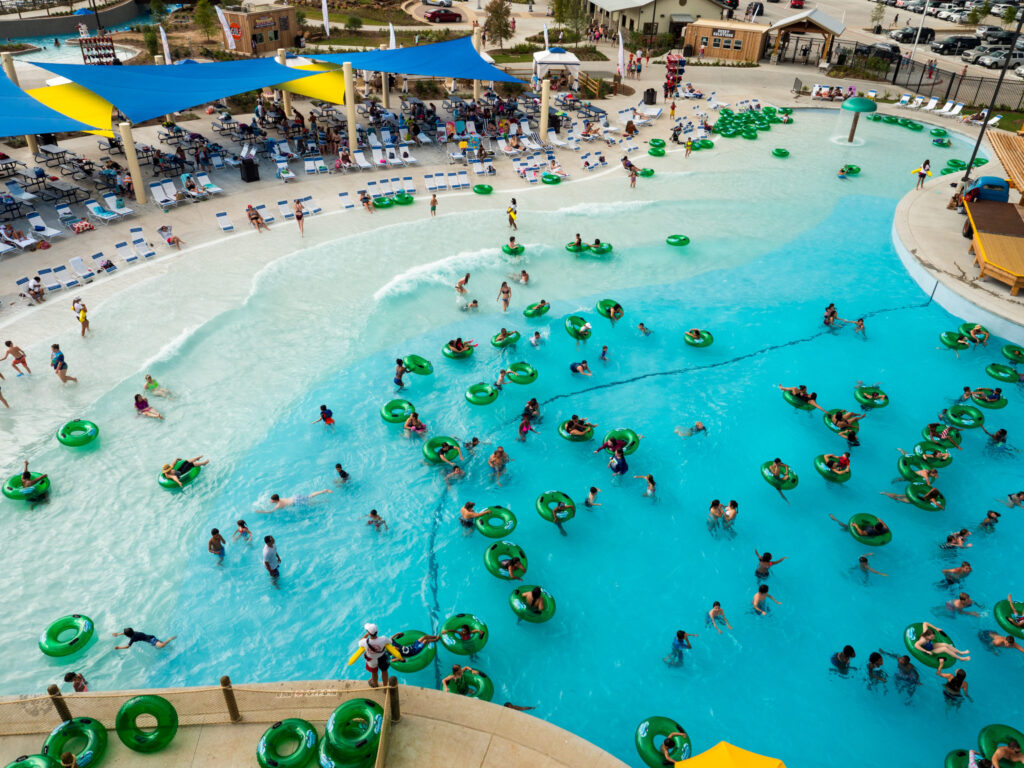 An elevated view of a crowded, sprawling wave pool at a water park. Many people are floating on green inner tubes, while others are wading or swimming in the bright blue water.