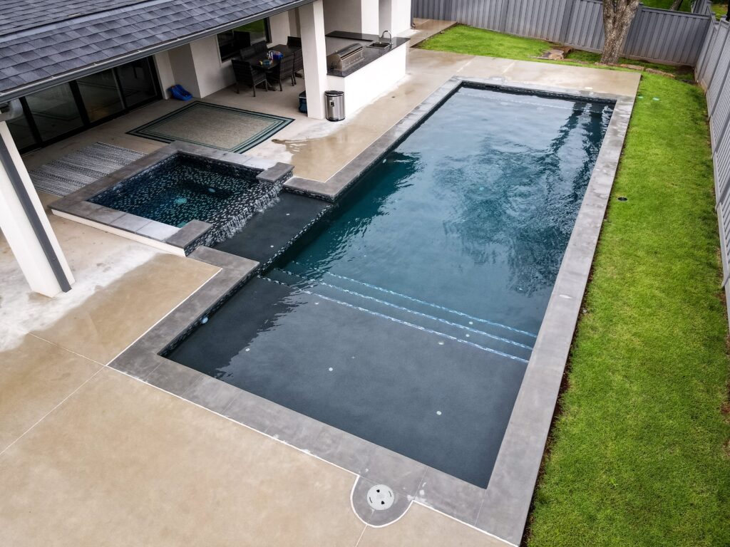 Rectangular in-ground pool adjacent to a contemporary home, surrounded by lush green grass.