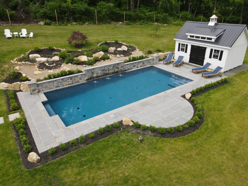 A rectangular swimming pool with blue water and stone coping sits in a lush green lawn, flanked by a stone retaining wall with water features on one side and a white shed with a black roof and four blue lounge chairs on the other.