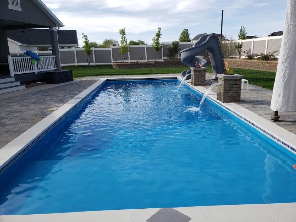 A rectangular backyard swimming pool with a vibrant blue finish, featuring two stone pillars with fire bowls and water features, a grey slide, and paved surrounding patio area.