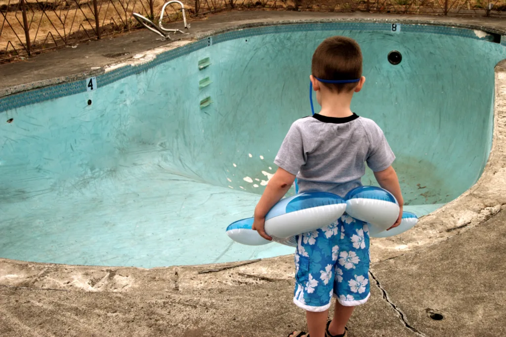 Small boy wearing a floaty and goggles stares at an empty swimming pool.