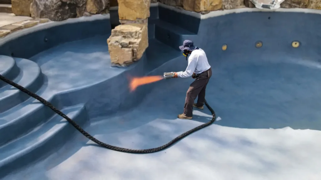 A construction worker wearing a hat and respirator mask is refinishing the interior of a large, empty, in-ground swimming pool. He is using a specialized spray gun to apply a new, blue finish to the curved steps and main basin.