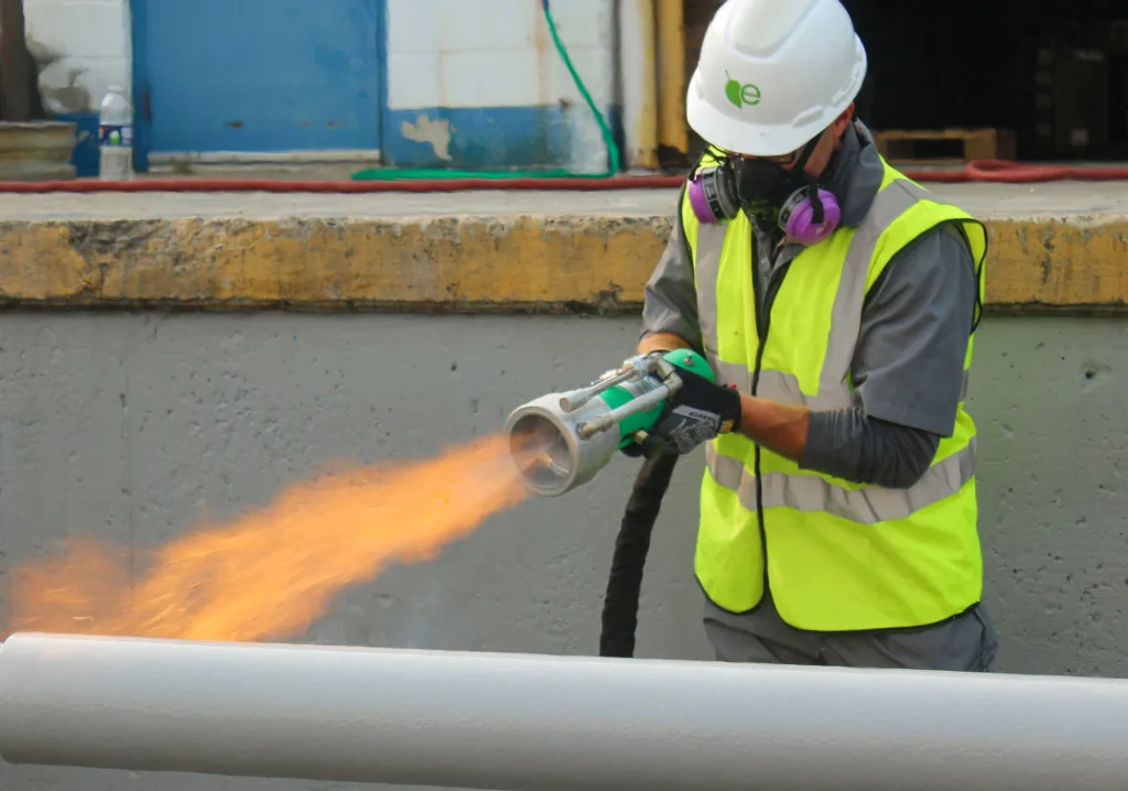 A worker wearing a white hard hat, respirator, and high-visibility vest uses a flame tool to apply a flame coating on a large, gray pipe in an industrial setting. The tool emits a bright orange flame directed at the pipe's surface.