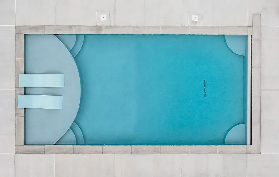 An overhead view of a rectangular swimming pool with a bright blue finish. The pool has a large, rounded section with steps and two submerged benches on the left side. The corners on the right side of the pool are rounded and also feature submerged benches. The pool is surrounded by a gray tile border and a light-colored tiled deck.