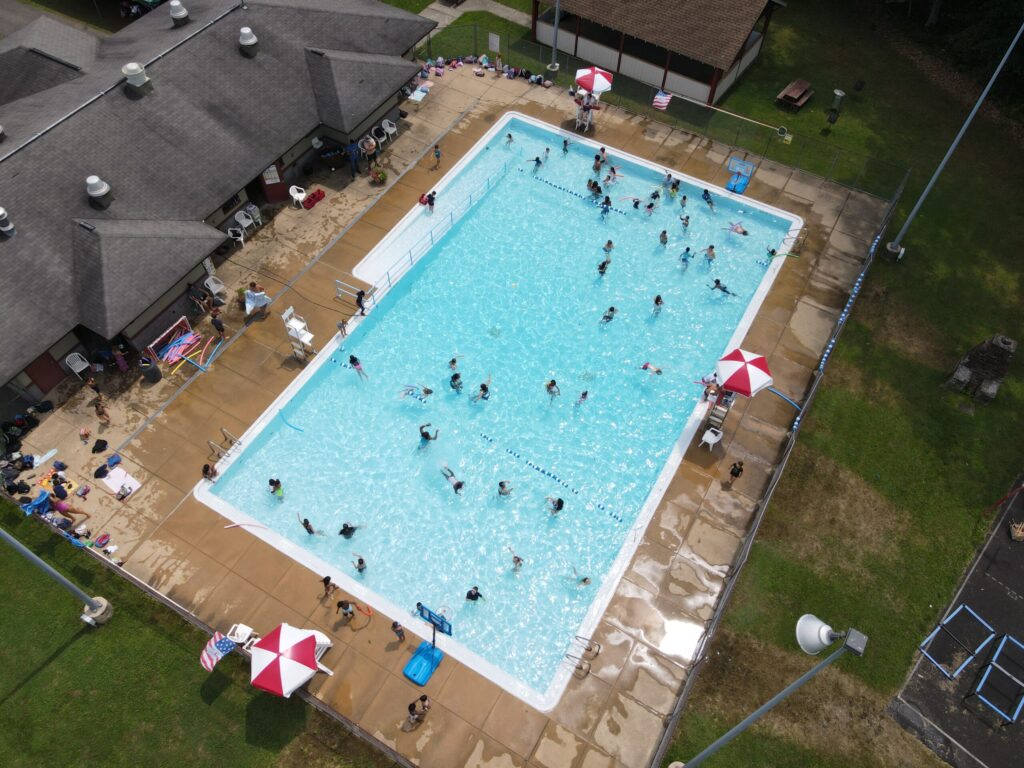 An aerial shot of a busy public swimming pool. The pool is rectangular with a light blue finish, and many people are swimming and playing in the water. Several people are sitting in chairs on the concrete deck, which has three red and white umbrellas.
