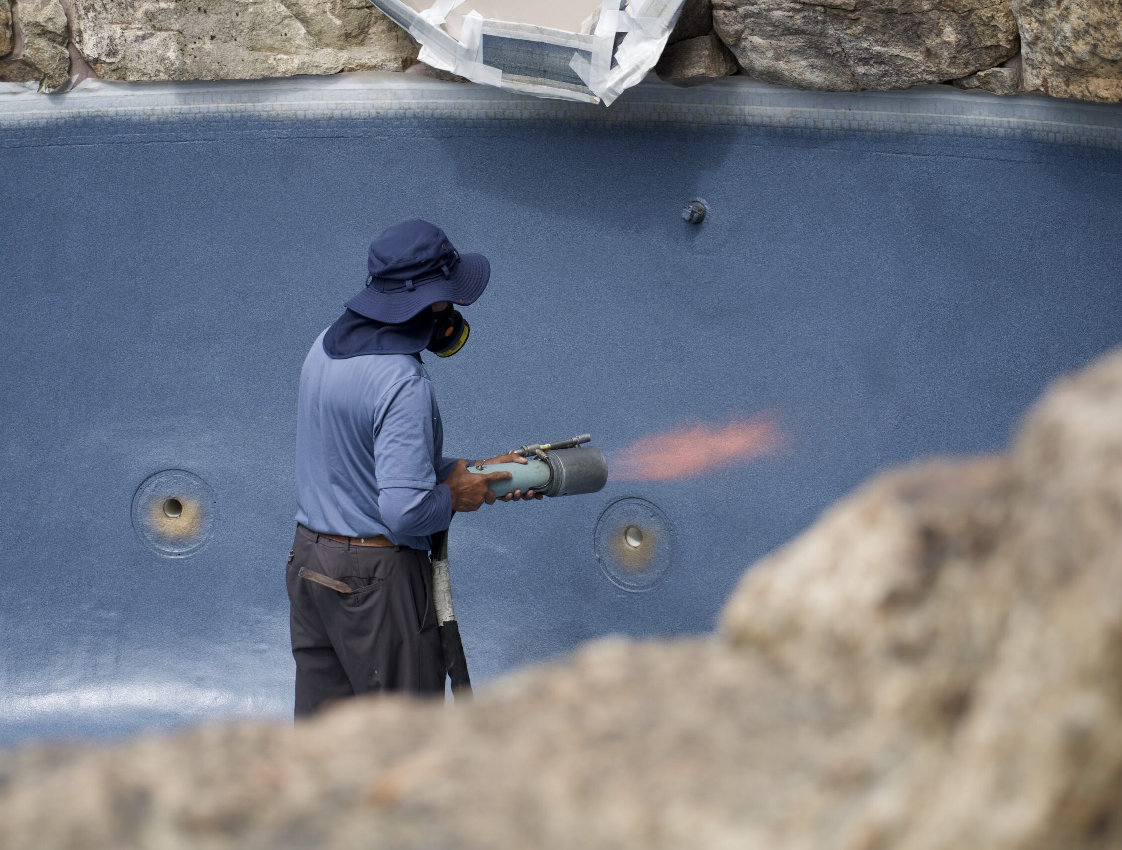 A side view shows a worker standing inside an empty swimming pool. The worker is holding a spray gun with a long hose, applying a blue coating to the pool wall. An orange flame is coming from the gun's nozzle, indicating an application process. The pool's surface is a dark blue, and the surrounding deck is composed of large, natural-colored rocks.