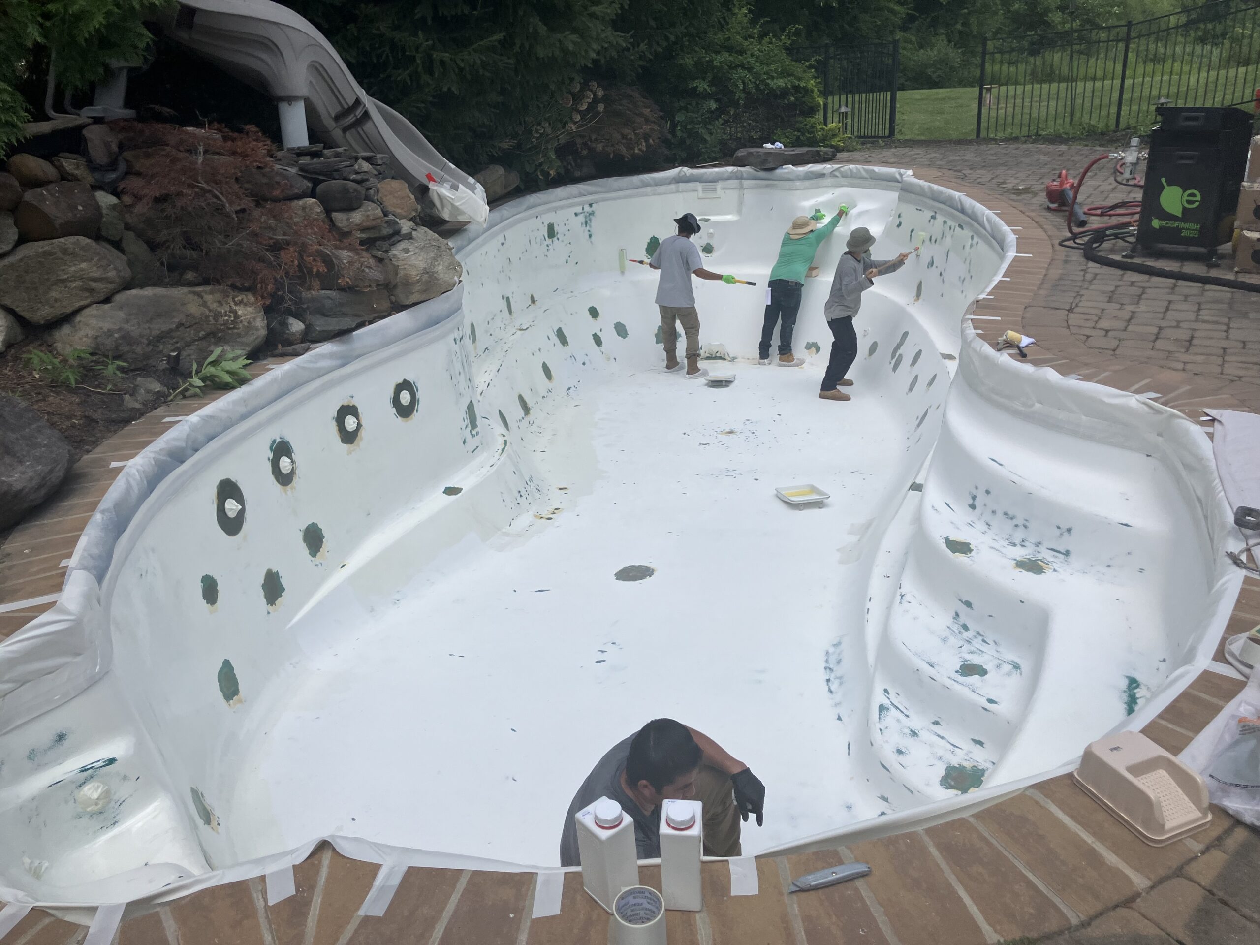 An image shows four workers engaged in renovating a kidney-shaped, in-ground swimming pool. The pool's interior is stripped bare, revealing its white fiberglass shell, which has some patches of green and brown residue. The pool deck is made of reddish-brown pavers.