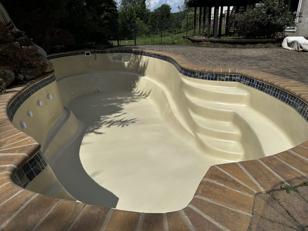 An in-ground, kidney-shaped swimming pool is shown from a high angle. The pool is empty and has a smooth, tan-colored finish. A set of wide steps is on the right side of the pool. The pool's edge is surrounded by a brown brick coping with a row of dark blue and white mosaic tiles just below it. The deck around the pool is made of red pavers.