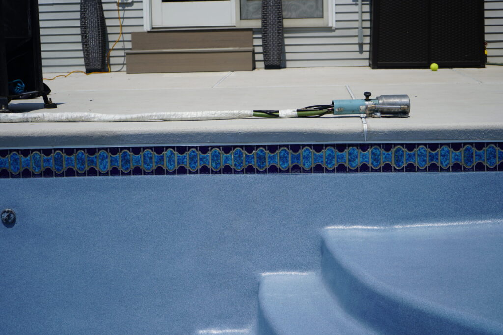 An eye-level, medium shot shows a swimming pool with an ecofinish coating in a blue lagoon color, tile, coping, concrete deck, stairs, a door, windows, siding, and an ecofinish applicator gun with a hose and a yellow tennis ball on a sunny day.